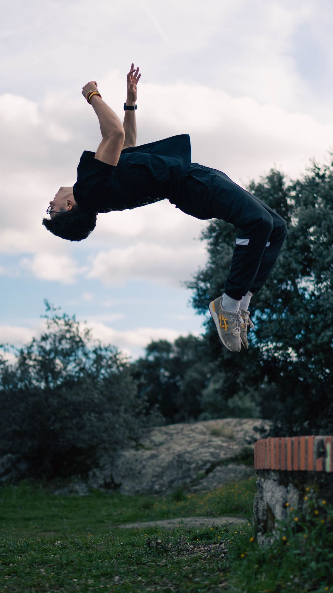 An energetic man performs a backflip in a natural outdoor environment with trees and clear skies.
