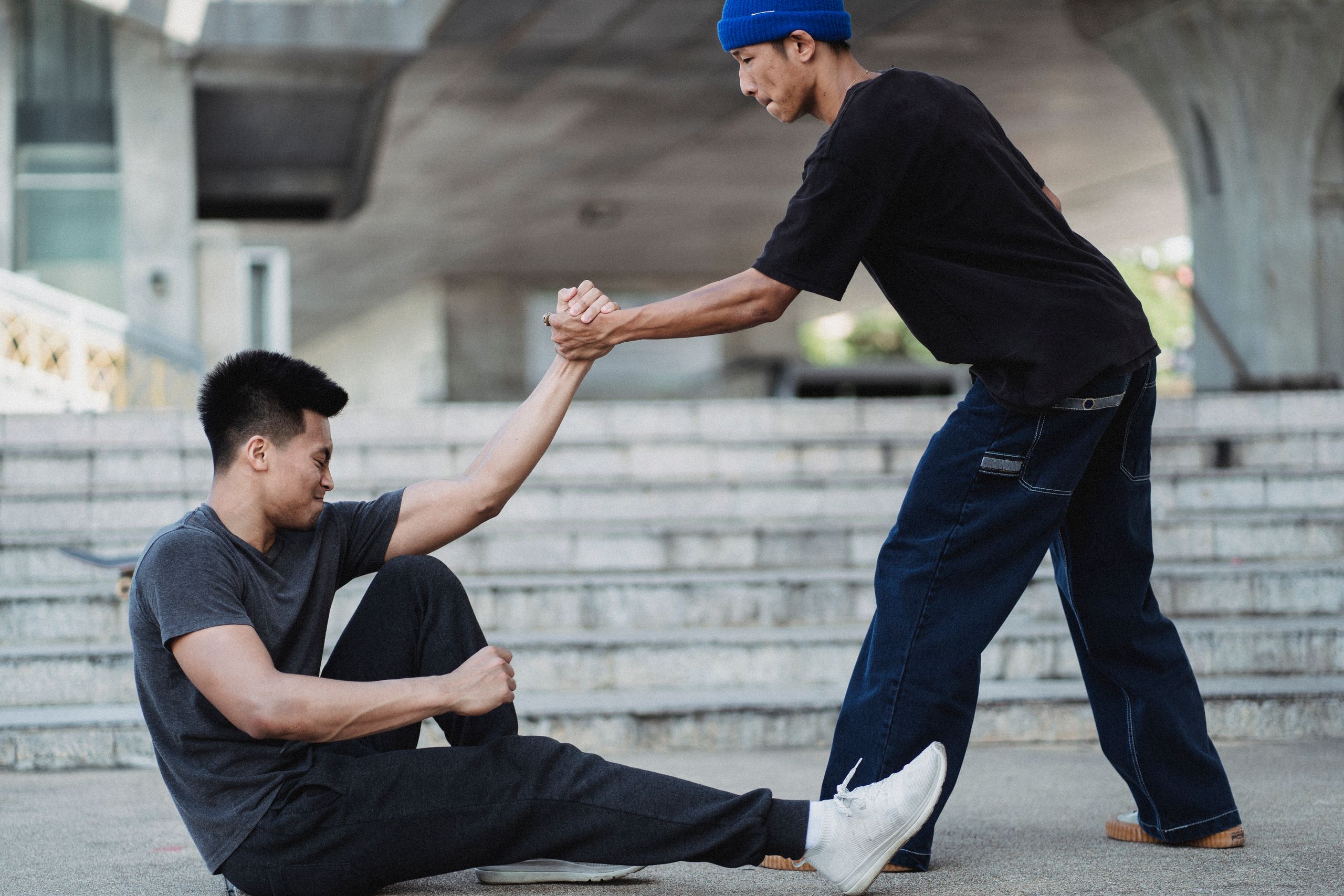 Side view Asian man in street wear holding friends hand and helping to get up from ground on sunny street