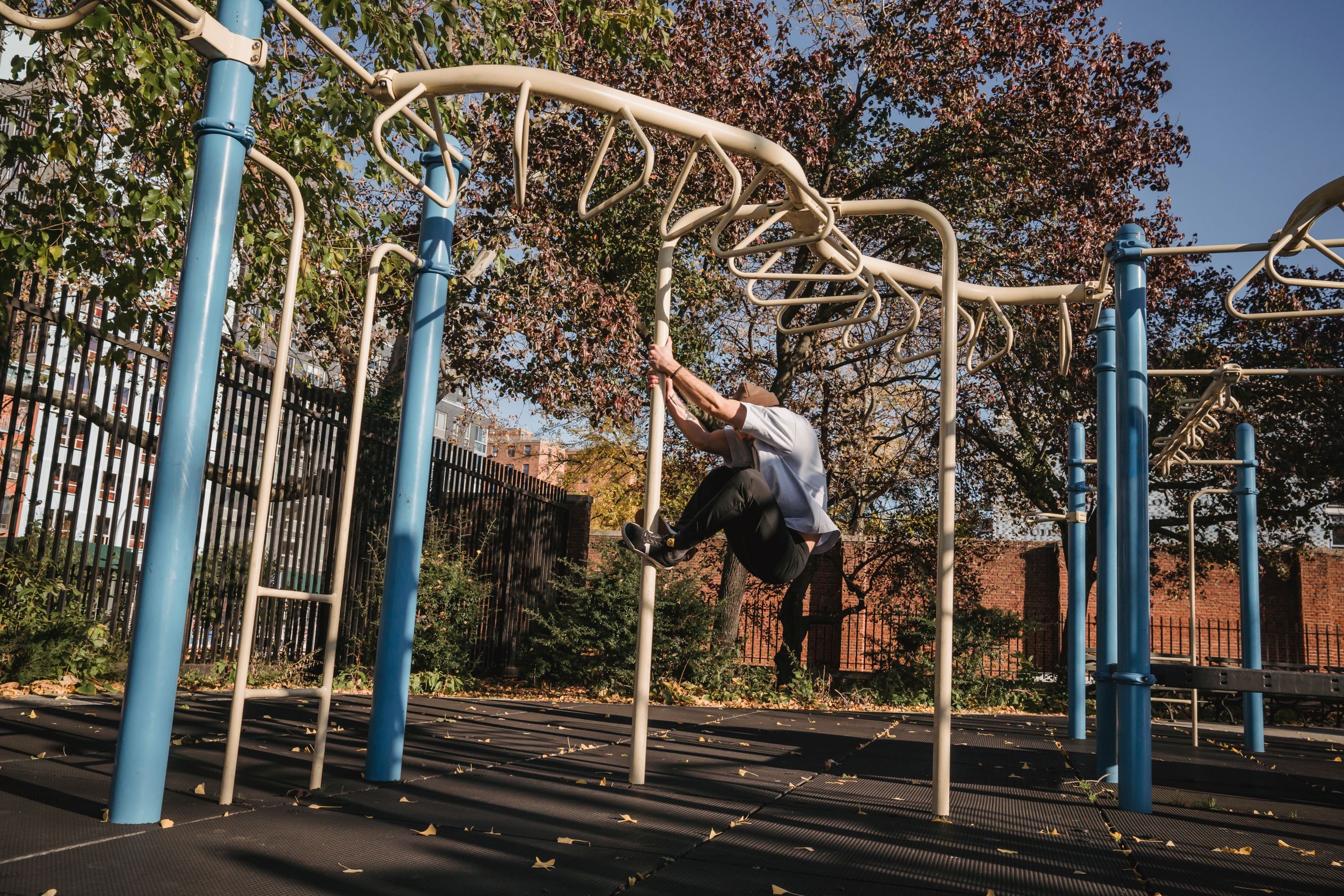 Side view full length of anonymous man hanging on pole in park near metal turnstile in sunny autumn day in street near trees and fence under blue cloudless sky